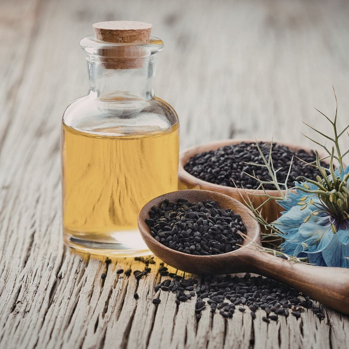 Glass bottle of golden black seed oil placed on a rustic wooden surface, with wooden spoons filled with black cumin (Nigella sativa) seeds and a blue Nigella flower beside it.
