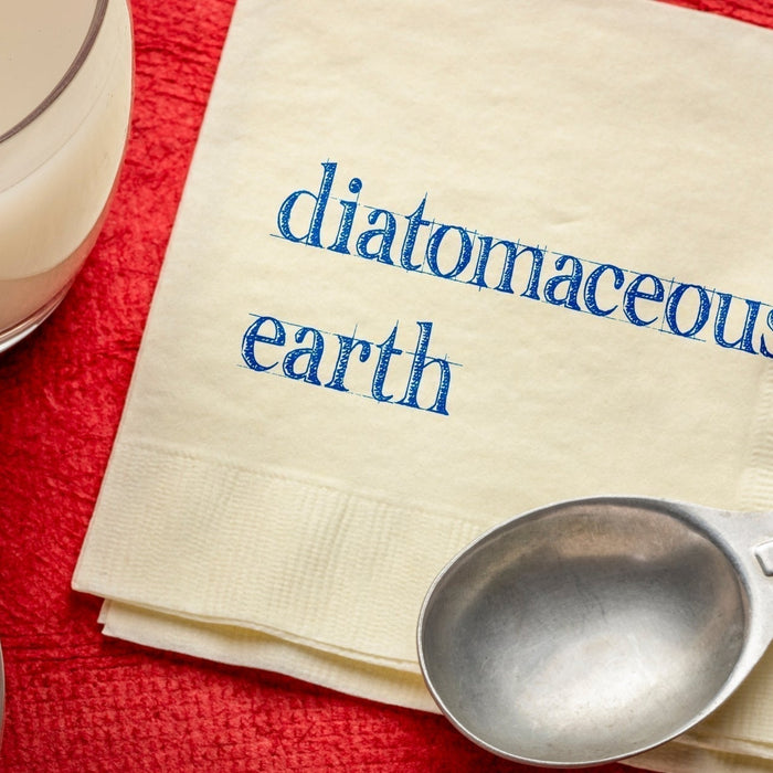 A top-down view of Diatomaceous Earth powder, a measuring tablespoon, and a glass of water on a red surface, with the words "diatomaceous earth" written on a napkin.