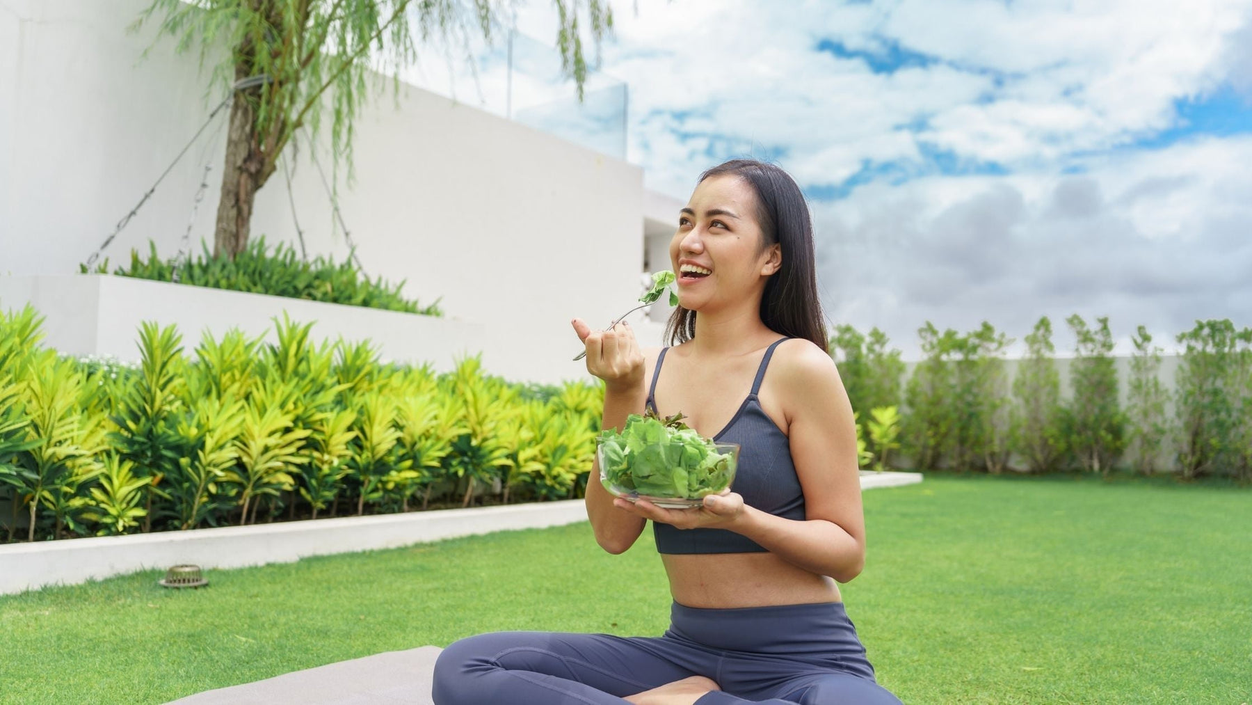 A happy woman sitting on a yoga mat outdoors eating a fresh green salad to improve metabolic health and energy.