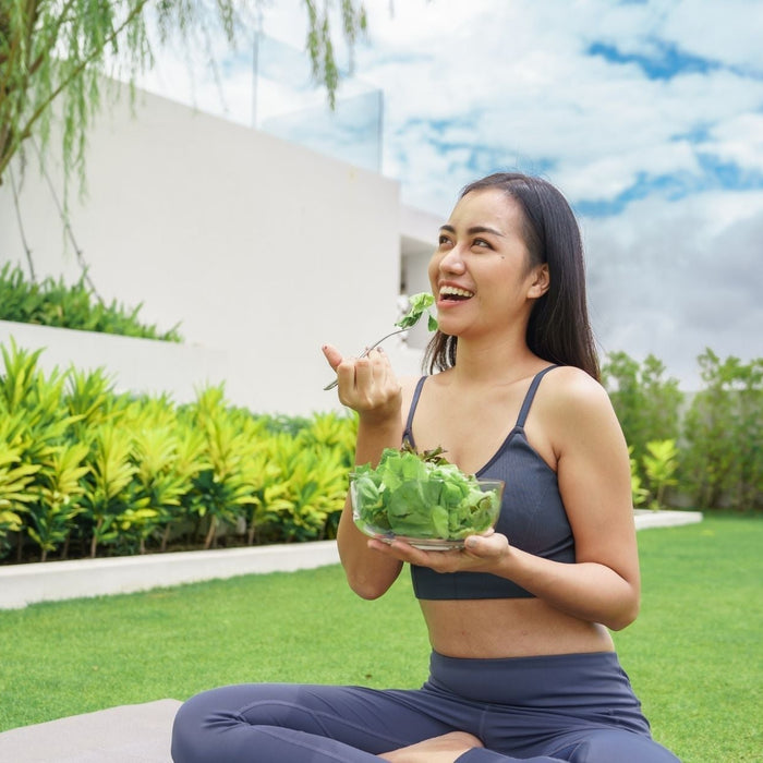 A happy woman sitting on a yoga mat outdoors eating a fresh green salad to improve metabolic health and energy.