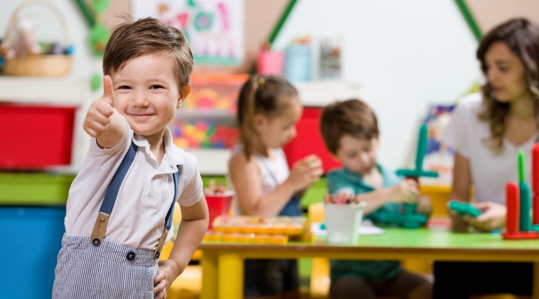 Happy preschool boy giving a thumbs up in a classroom with kids doing sensory activities in the background