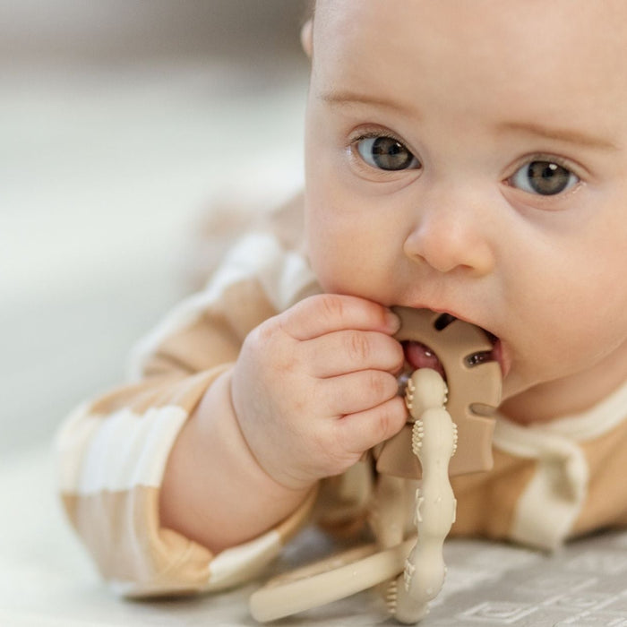 A close-up shot of a baby in a tan and white striped onesie lying down and chewing on a light brown, textured silicone teething ring to soothe their gums.