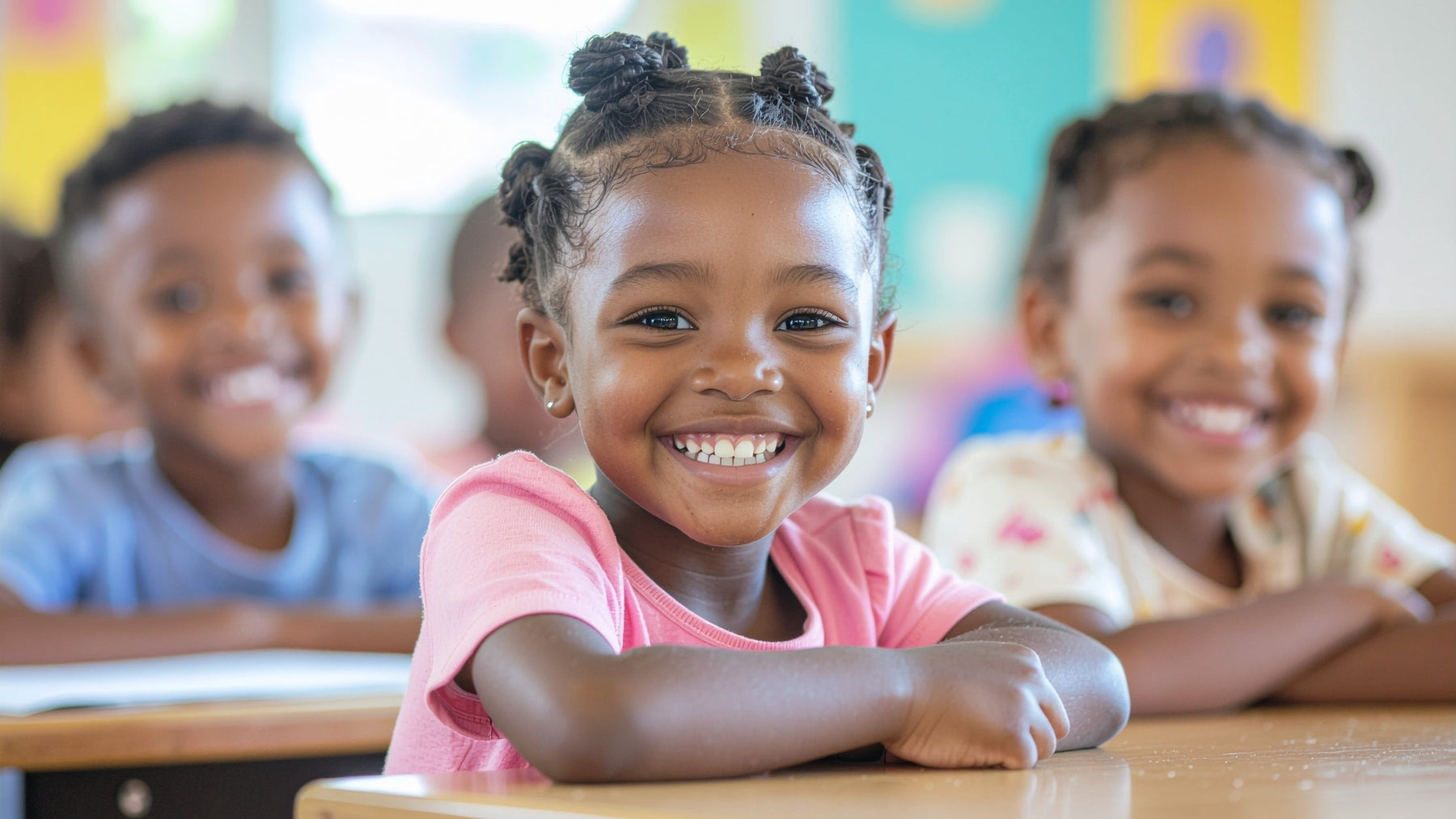 Three young children sitting at desks in a classroom, smiling broadly, looking healthy and happy.