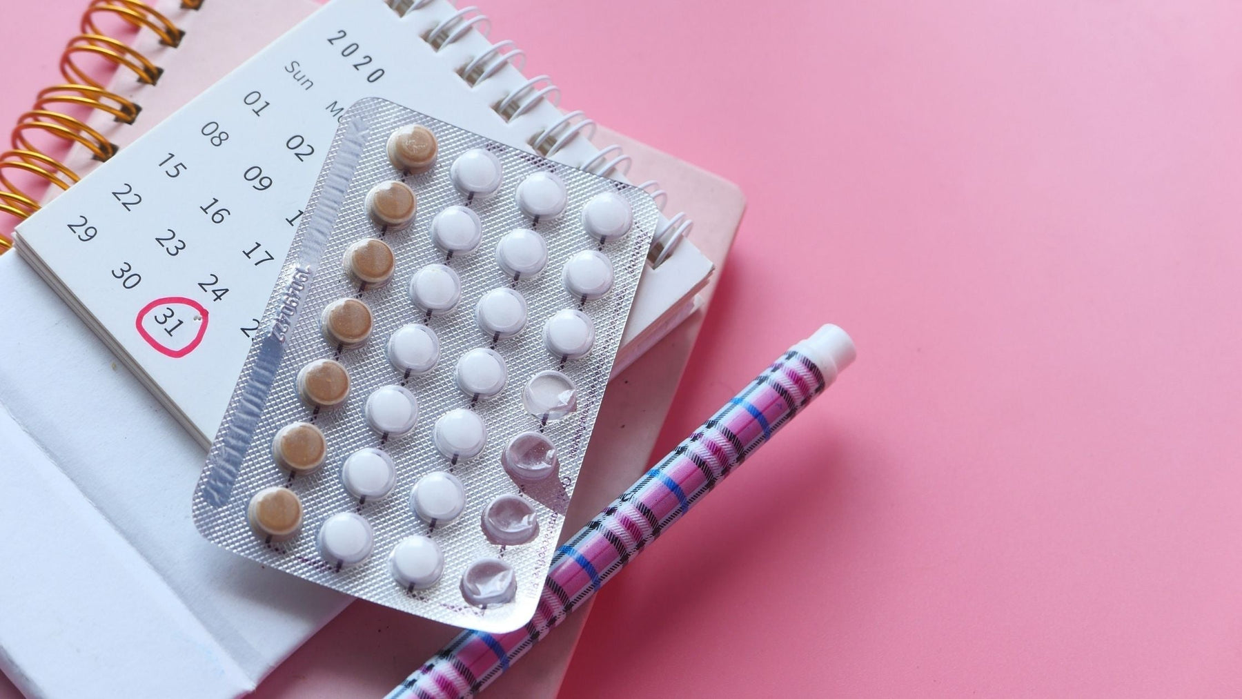 Pack of birth control pills sitting on a calendar planner with a pen on a pink background.