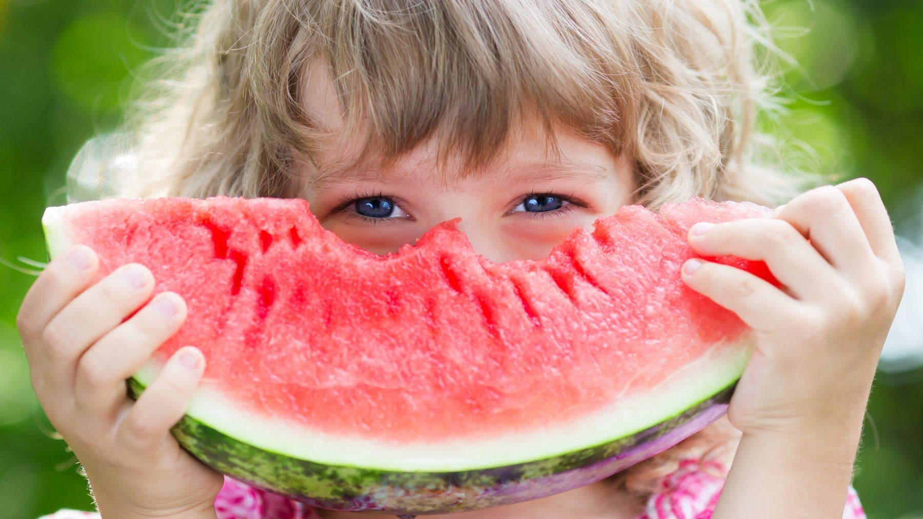 A young child holding a large slice of fresh, ripe watermelon over their face, highlighting healthy whole foods for kids.