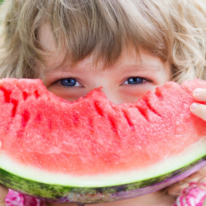 A young child holding a large slice of fresh, ripe watermelon over their face, highlighting healthy whole foods for kids.