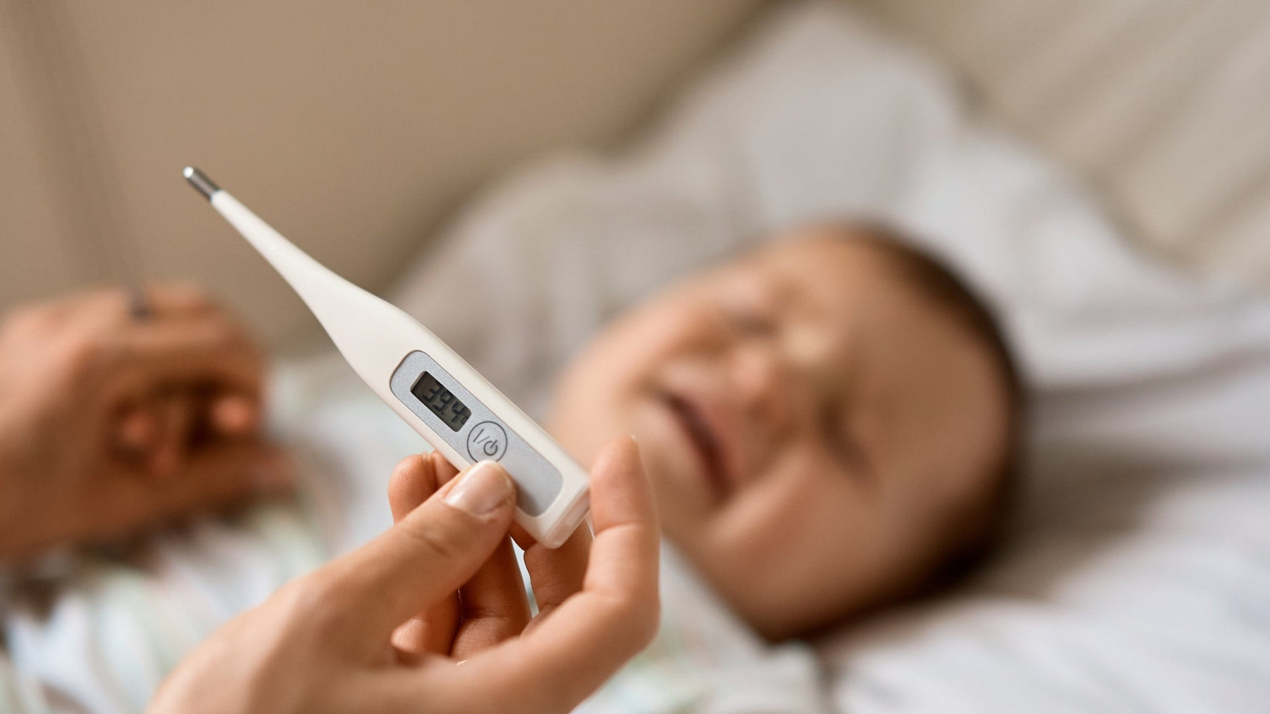 A close-up of an adult's hand holding a white digital thermometer displaying a high fever reading of $39.4\text{ }^\circ\text{C}$, with a crying, fussing baby lying down in the blurred, soft-focus background.