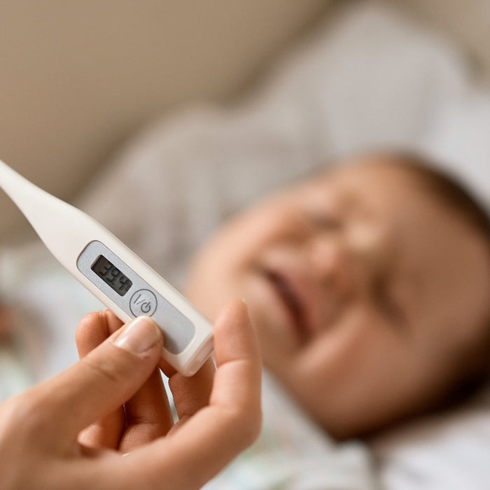 A close-up of an adult's hand holding a white digital thermometer displaying a high fever reading of $39.4\text{ }^\circ\text{C}$, with a crying, fussing baby lying down in the blurred, soft-focus background.