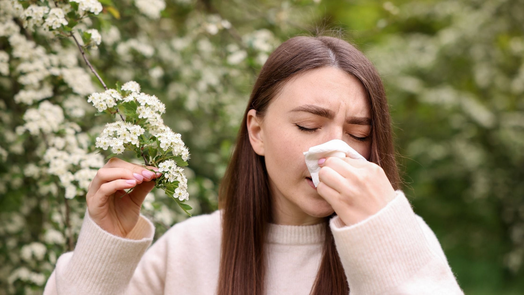 A woman sneezing into a tissue outdoors next to blooming white spring flowers, illustrating seasonal allergies and the body’s natural reaction to high pollen counts.