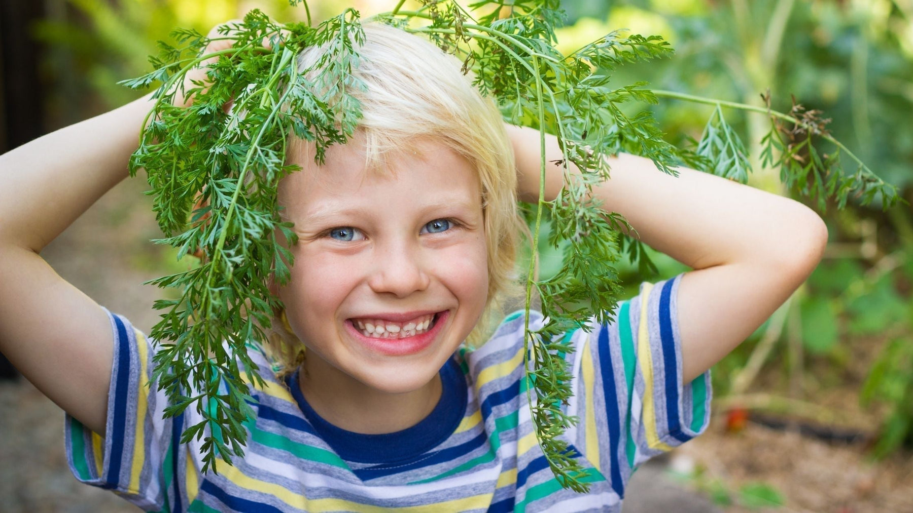 Happy, smiling young boy outdoors holding fresh carrot greens on his head, showcasing outdoor play and natural nutrition for child focus.