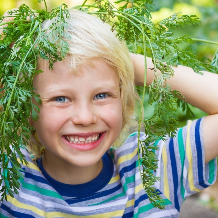 Happy, smiling young boy outdoors holding fresh carrot greens on his head, showcasing outdoor play and natural nutrition for child focus.