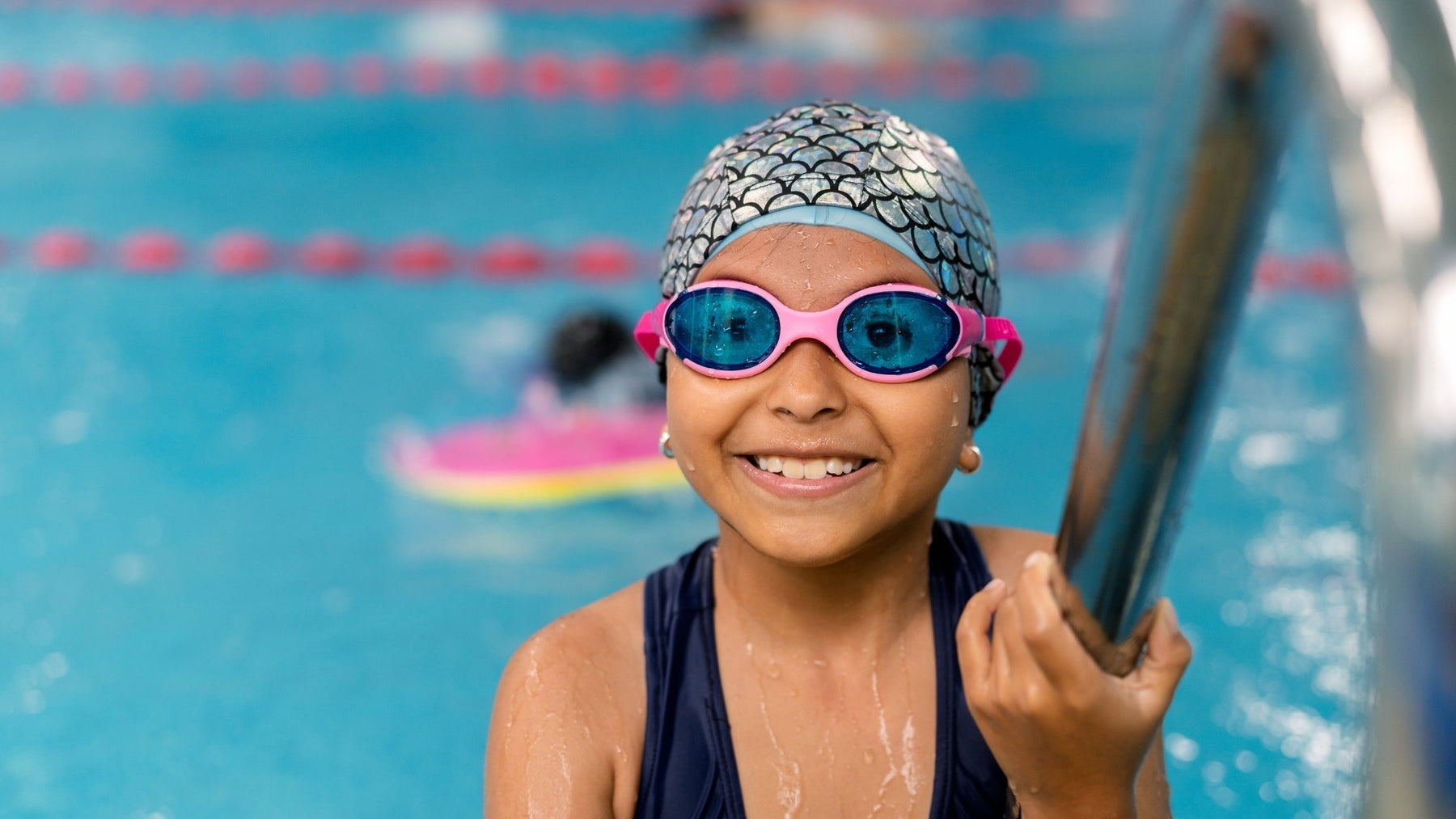 Young girl smiling in a swimming pool wearing pink goggles and a swim cap, demonstrating swimming as an effective individual after-school sport for ADHD support.