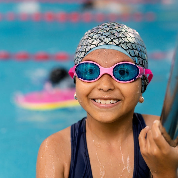 Young girl smiling in a swimming pool wearing pink goggles and a swim cap, demonstrating swimming as an effective individual after-school sport for ADHD support.