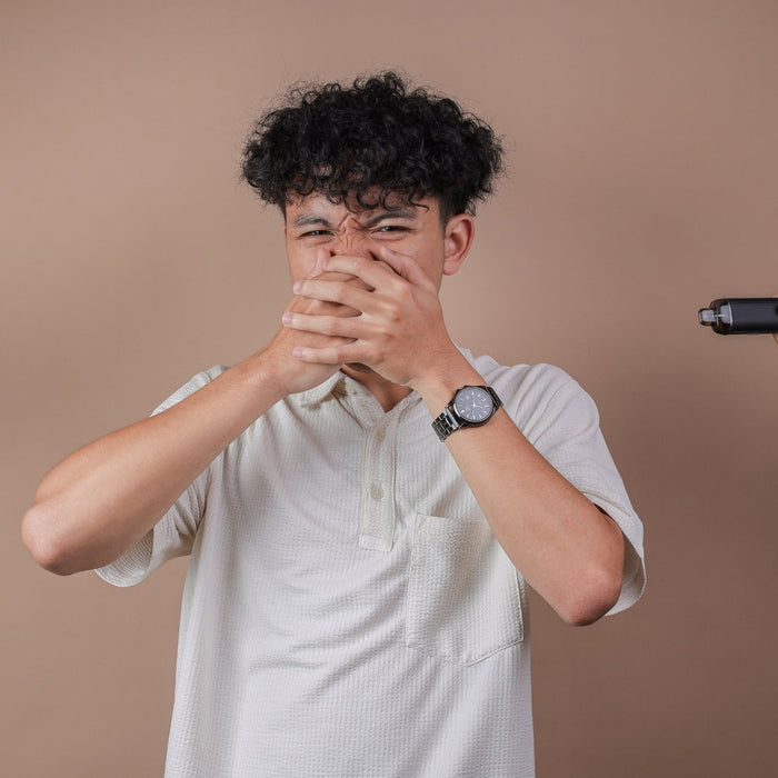 A young man covering his nose and mouth with both hands in refusal, while two hands extend from the sides of the frame offering him cigarettes on the left and a vape on the right.