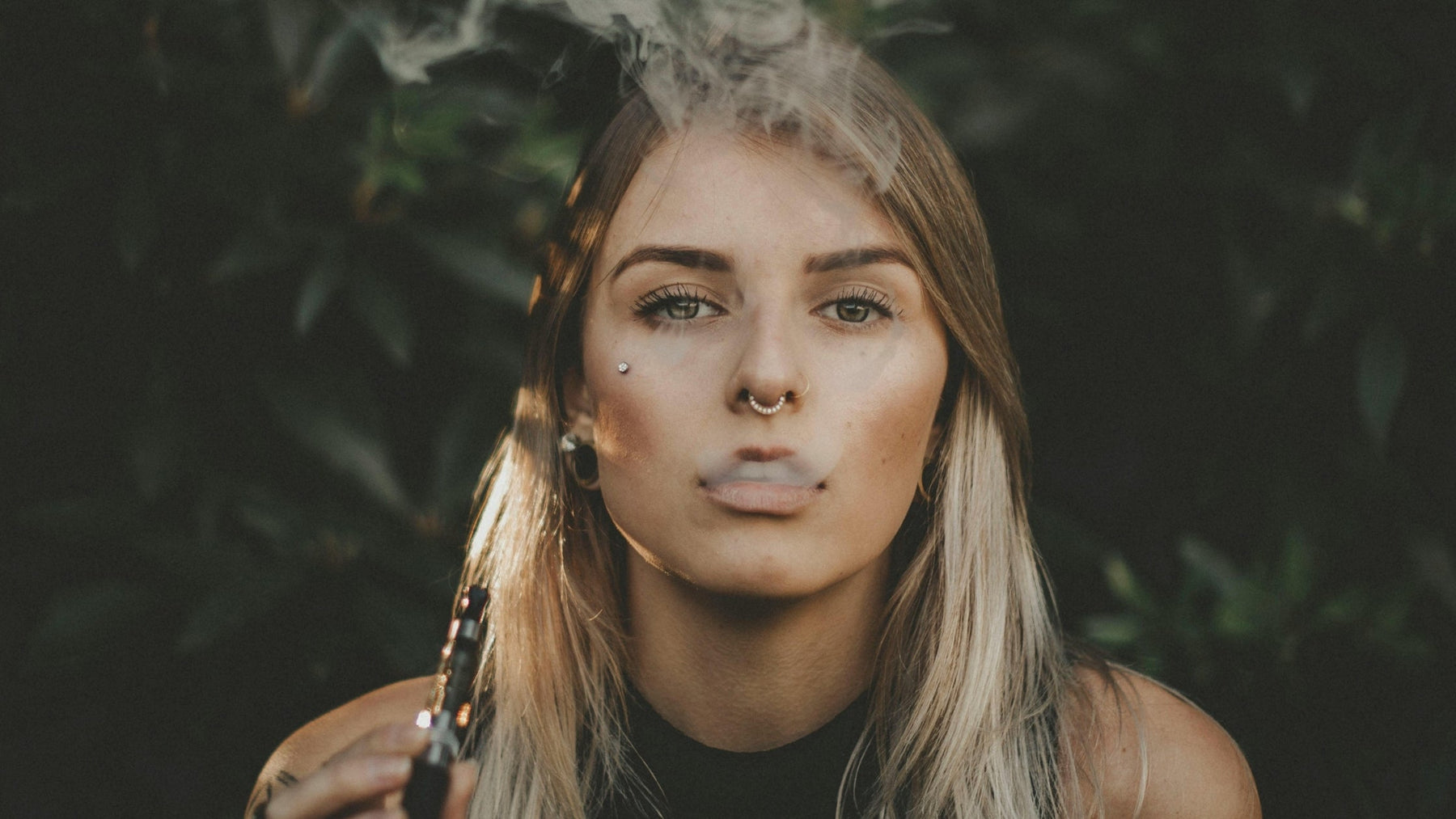 Young woman exhaling vape smoke outdoors while holding a vape device, illustrating stress-induced vaping cravings.