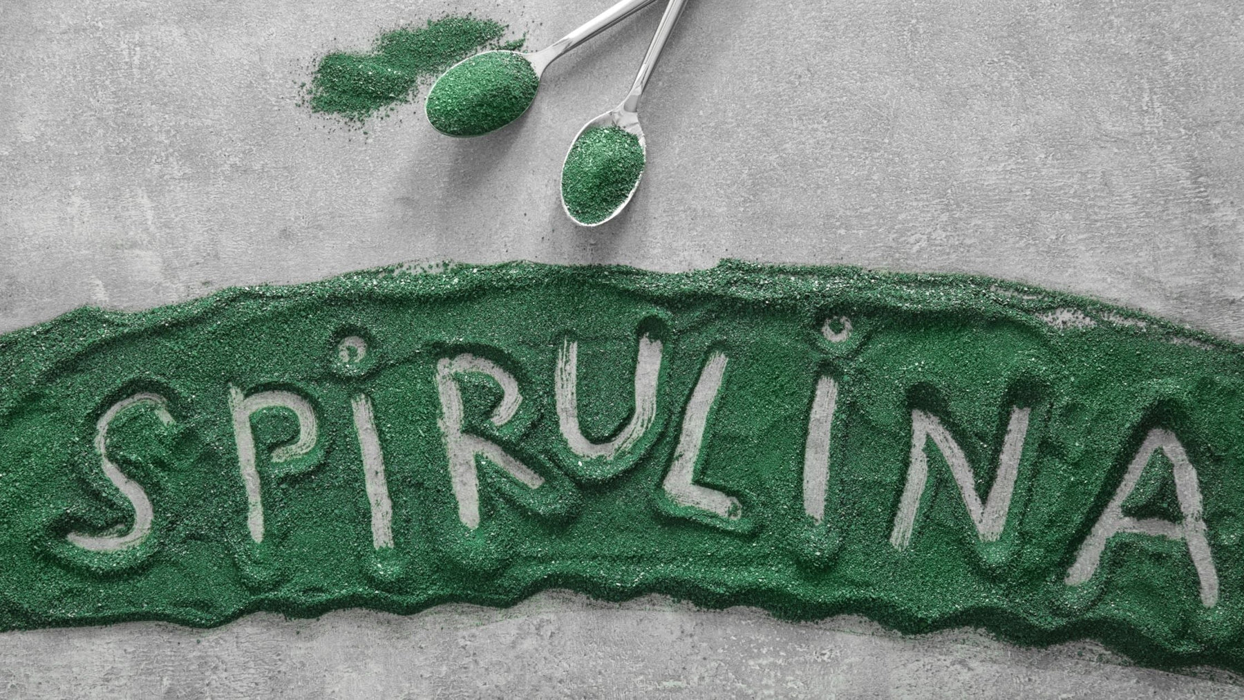 Green spirulina powder arranged on a grey surface with the word ‘spirulina’ written through the powder